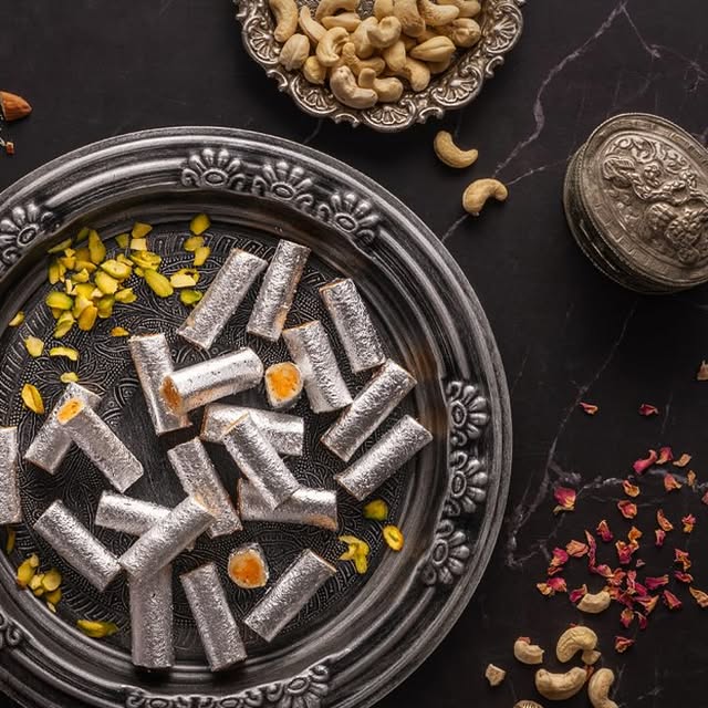 Silver-leaf kaju badam rolls arranged on a dark serving plate